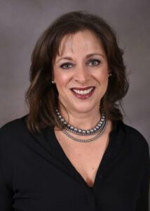 Headshot of Beth Calhoun-Leipold wearing a black v-neck blouse, with silver bead earrings, a silver beaded necklace and two silver box chain necklaces. She is the owner and managing partner of Work Horse Integrations, a company that can connect software systems for your business.