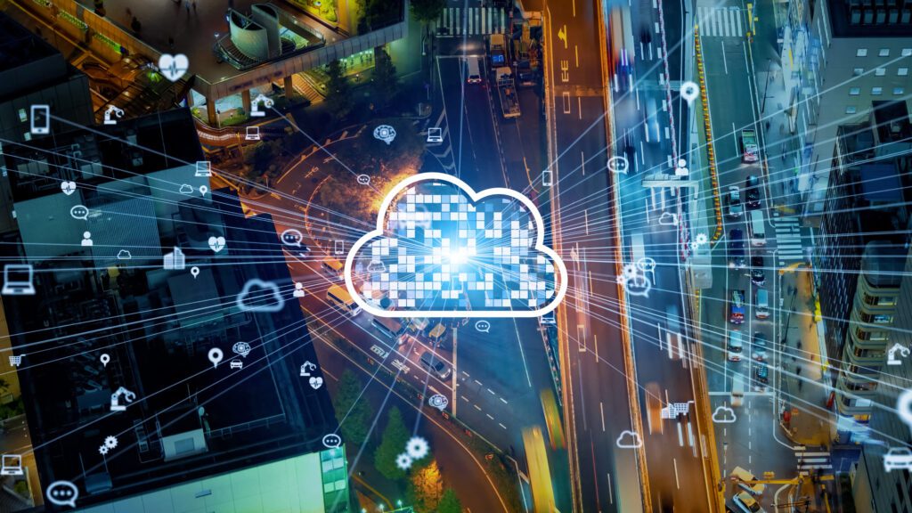 An illustration of a white and blue digital cloud in the sky overlooks a photo of a downtown city street with tall buildings at night. The interior of the cloud is made of a grid of small squares. The cloud has a burst of thin white lines shooting out from it, leading to various icons like a laptop and smart phone suggestive of our software integration platform.