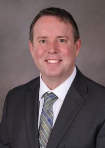 Headshot of Erich Leipold wearing a dark gray suit, white collared shirt and light gray tie with blue, purple, and yellow squares and diagonal white lines. Erich is Managing Partner & Senior Integration Architect of Work Horse Integrations, a company that can connect software systems for your business.